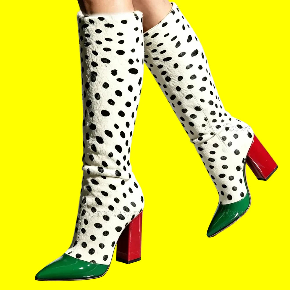 A pair of knee-high boots with a white faux-leather upper covered in black spots, featuring bright green pointed toes and red block heels, shown on a model against a light gray background.