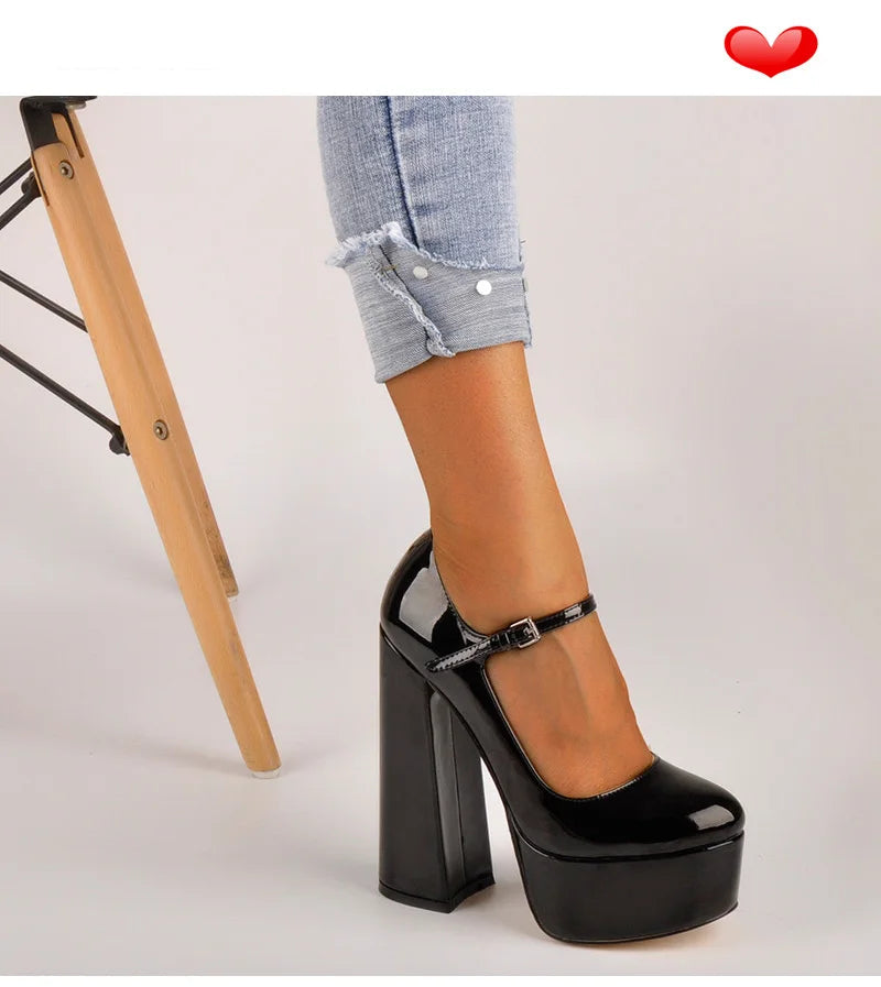 A person wearing a black patent leather high-heeled shoe with a platform sole and ankle strap, paired with light blue rolled-up jeans. A wooden chair leg is visible on the left against a light gray studio background.