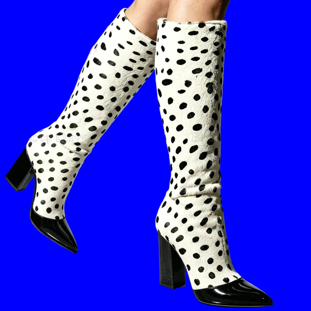 A pair of knee-high boots with a white faux-fur texture covered in black spots, featuring glossy dark red pointed toes and block heels, shown on a model against a light gray background.