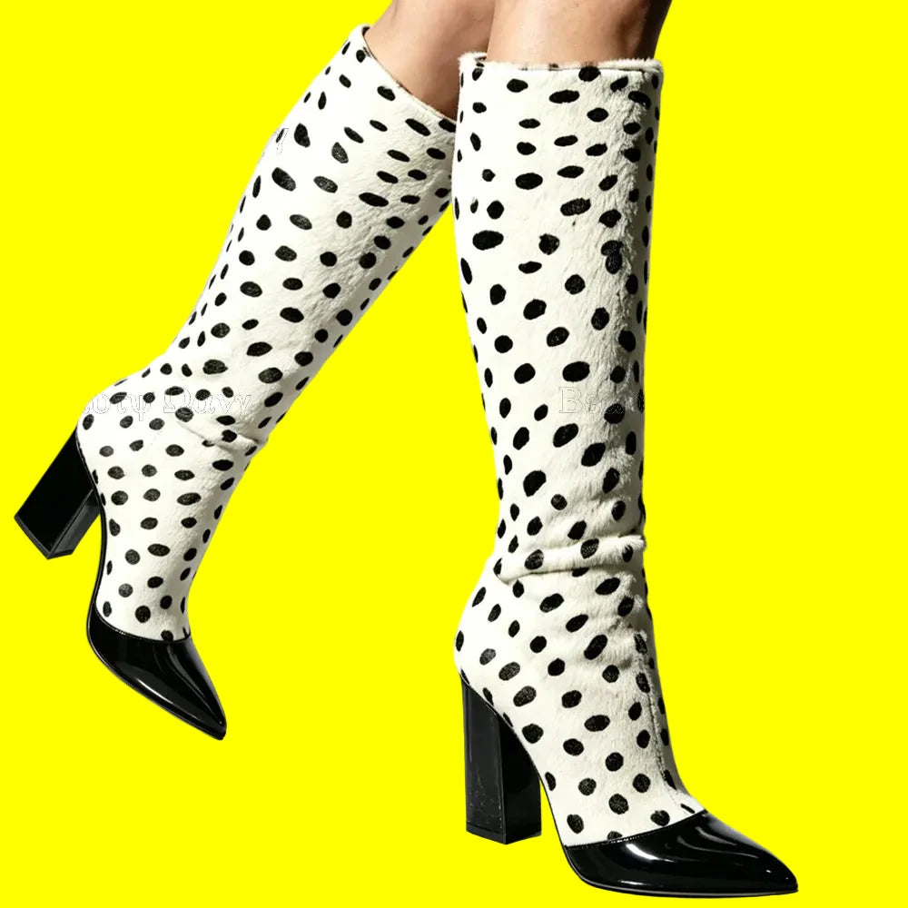 A pair of knee-high boots with a black and white dalmatian spot pattern and glossy black pointed toes and block heels, shown on a model's legs against a light gray studio background.