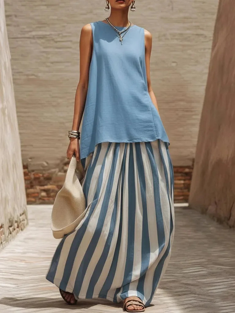 Woman wearing a blue sleeveless top and striped pants, holding a beige bag, against a stone wall background.