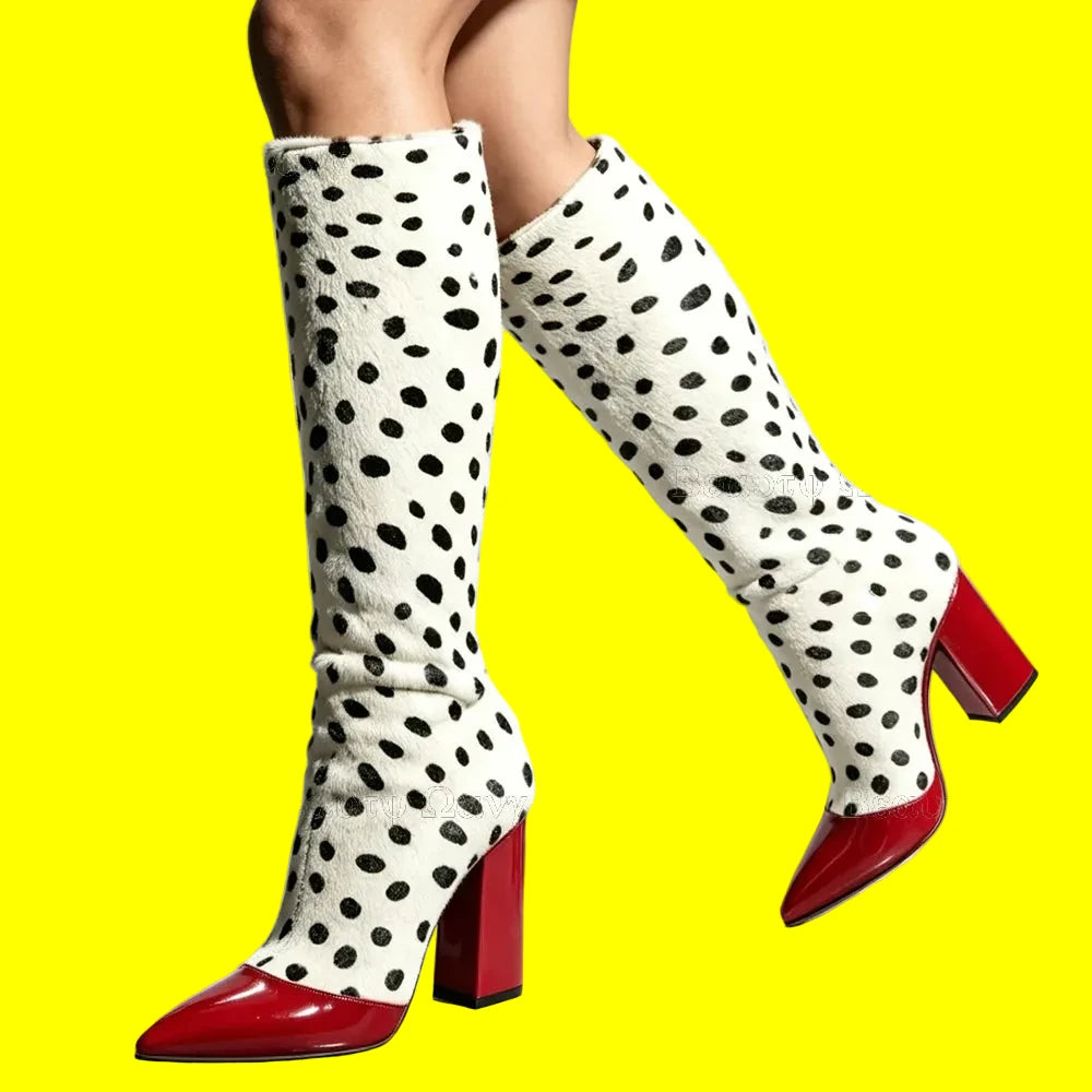 A pair of knee-high boots with a white faux-fur texture covered in black spots, featuring glossy red pointed toes and block heels, shown on a model's legs against a light gray background.