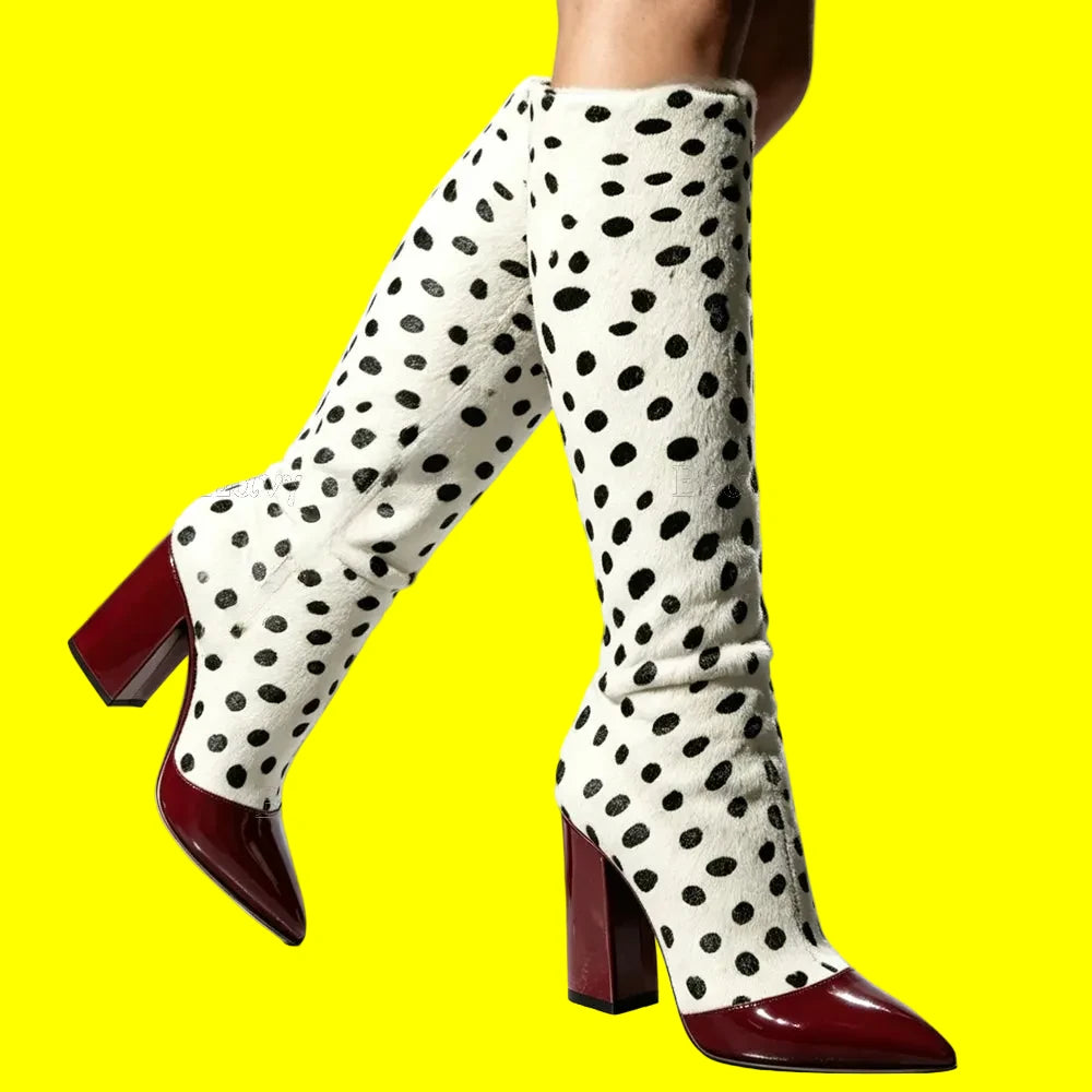 A pair of knee-high boots with a white faux-fur texture covered in black spots, featuring glossy dark red pointed toes and block heels, shown on a model against a light gray background.