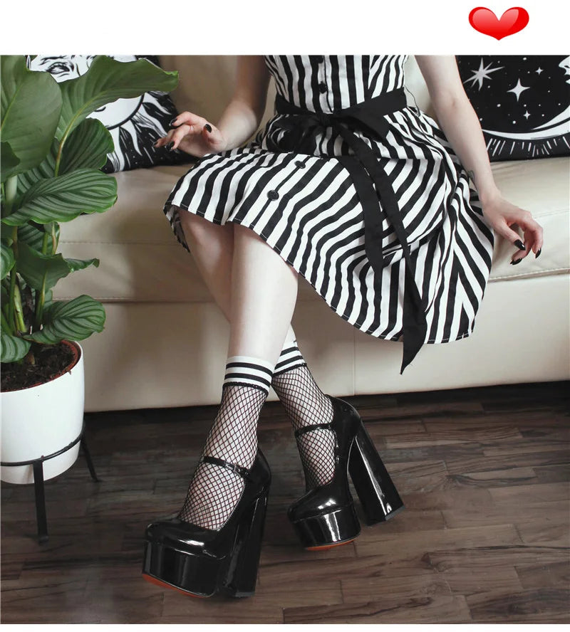 A person seated on a cream sofa wearing a black-and-white striped dress, striped fishnet socks, and glossy black platform heels with fishnet stockings. A potted plant and wood floor are visible, and a red heart icon appears in the top right corner.