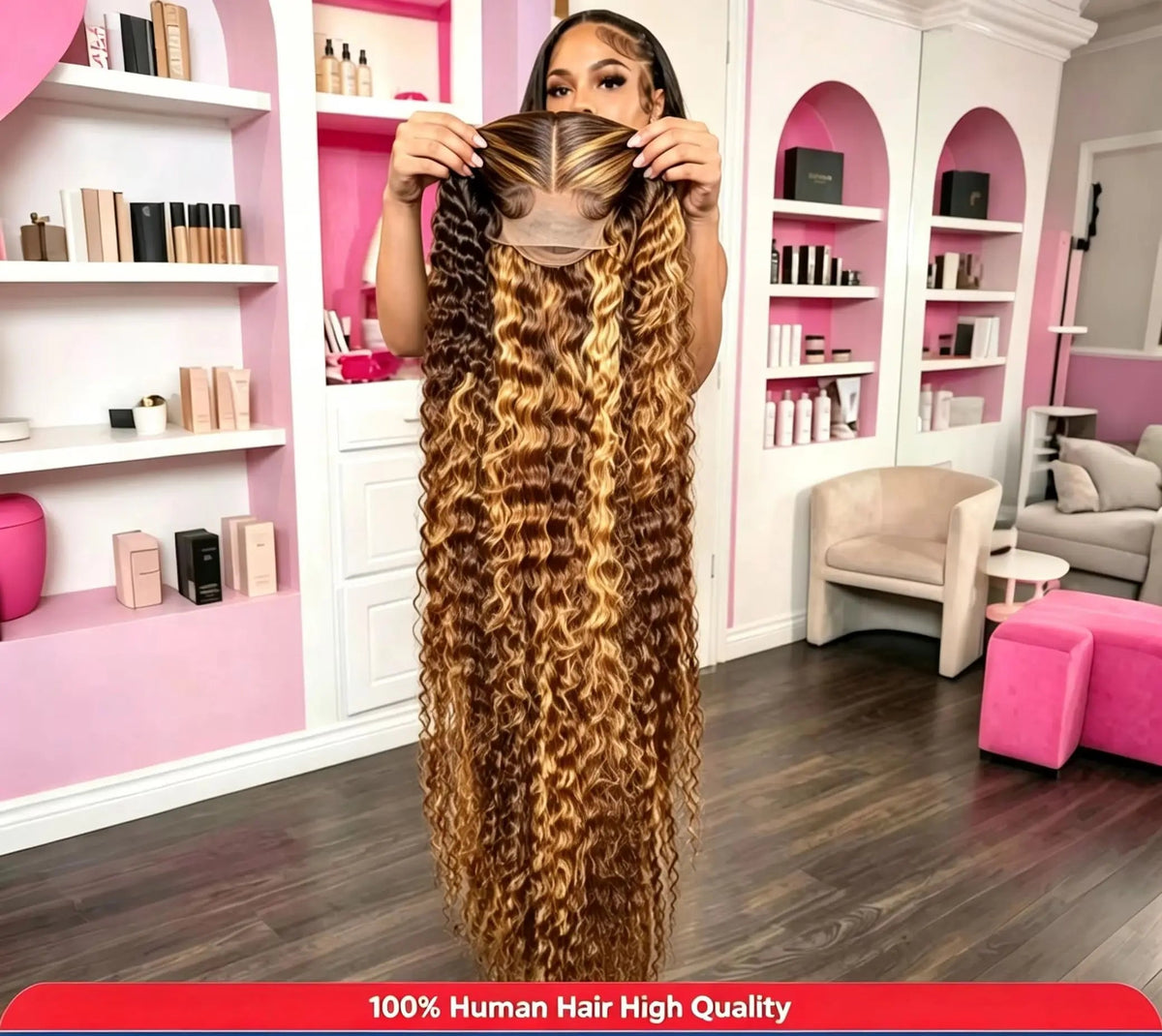 A woman in a salon-like room holds up a very long, wavy human hair extension piece with a dark brown to golden-brown ombre blend. The background shows pink and white shelves with hair products.