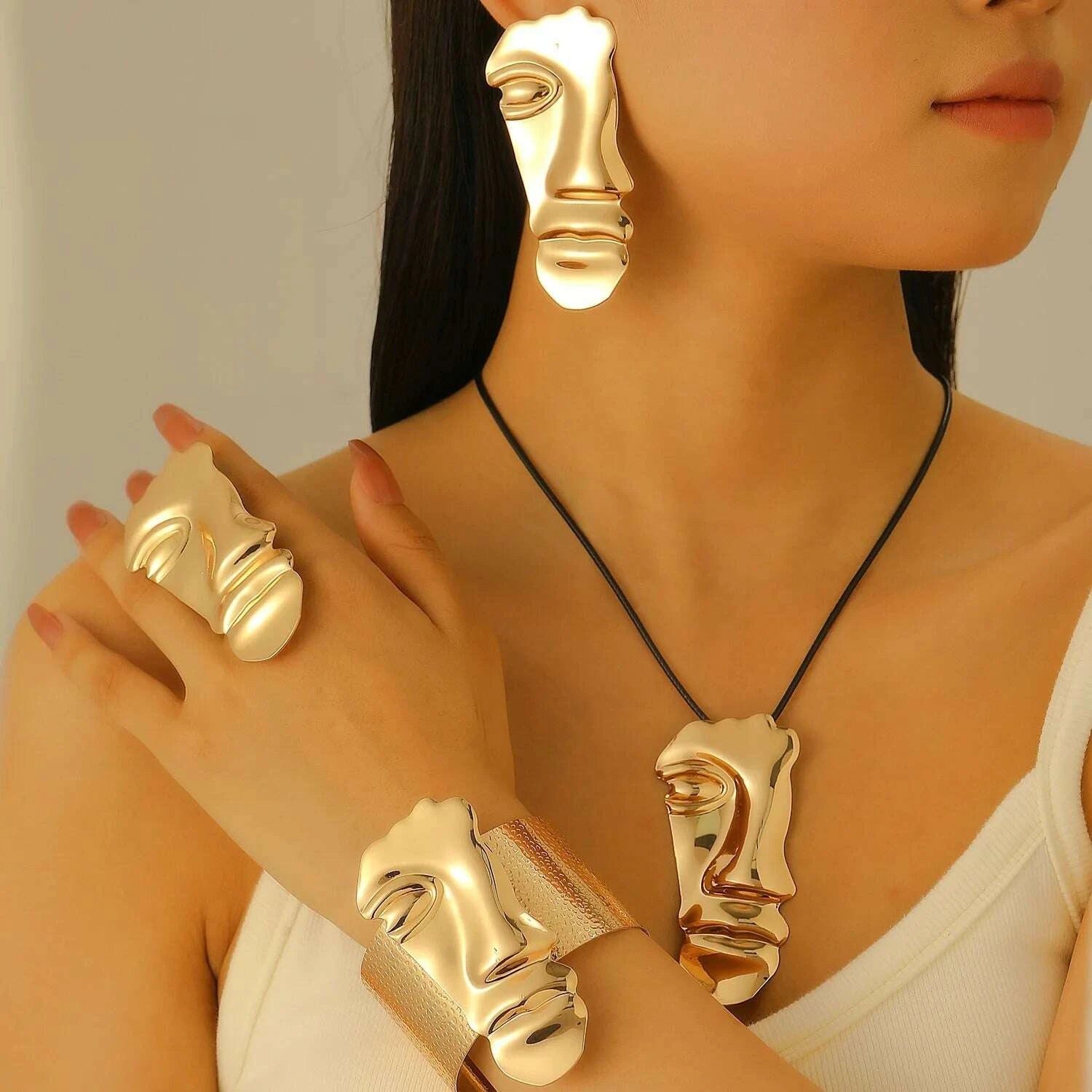 Woman wearing gold face-shaped jewelry including earrings, ring, and bracelet on a neutral background