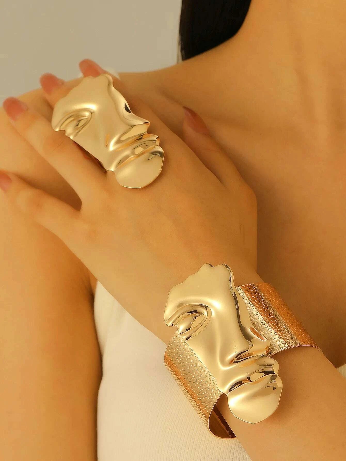 Gold face-shaped cuff bracelets on a person's wrist against a neutral background