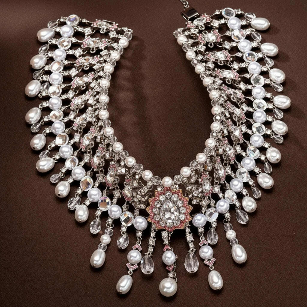 A highly detailed statement bib necklace featuring white freshwater pearls, clear teardrop crystals, and a central floral cluster with pink rhinestones on a silver-tone setting, displayed on a dark brown textured background.