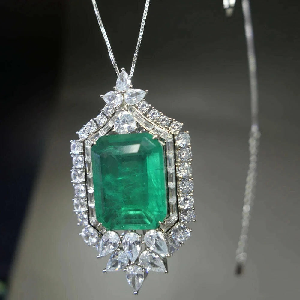 An ornate rectangular emerald-cut green gemstone pendant necklace set in silver-toned metal with surrounding clear stones, hanging against a dark gray background.
