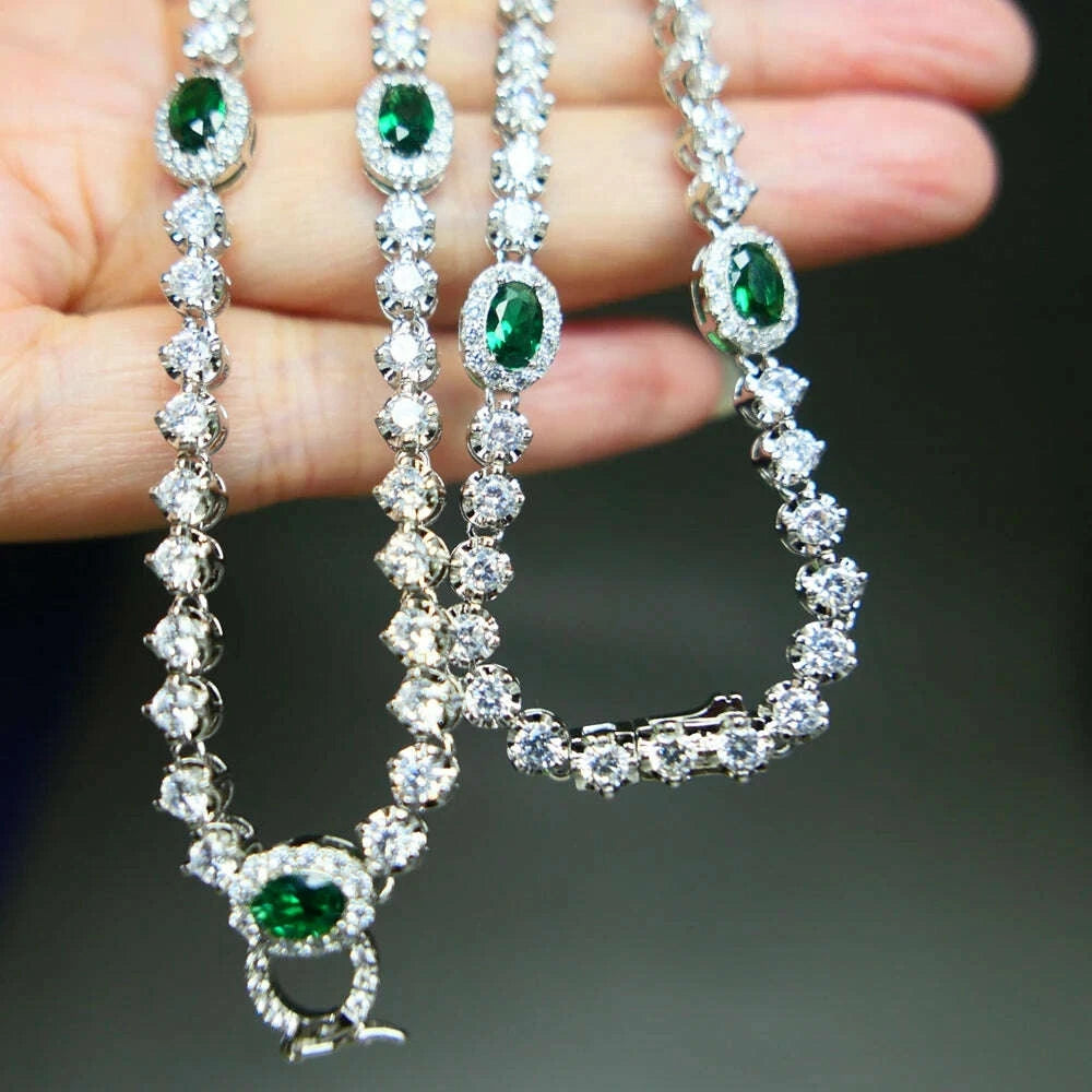 A close-up of a silver-toned necklace with oval green gemstones and clear stones, held in a person's hand against a dark blurred background.
