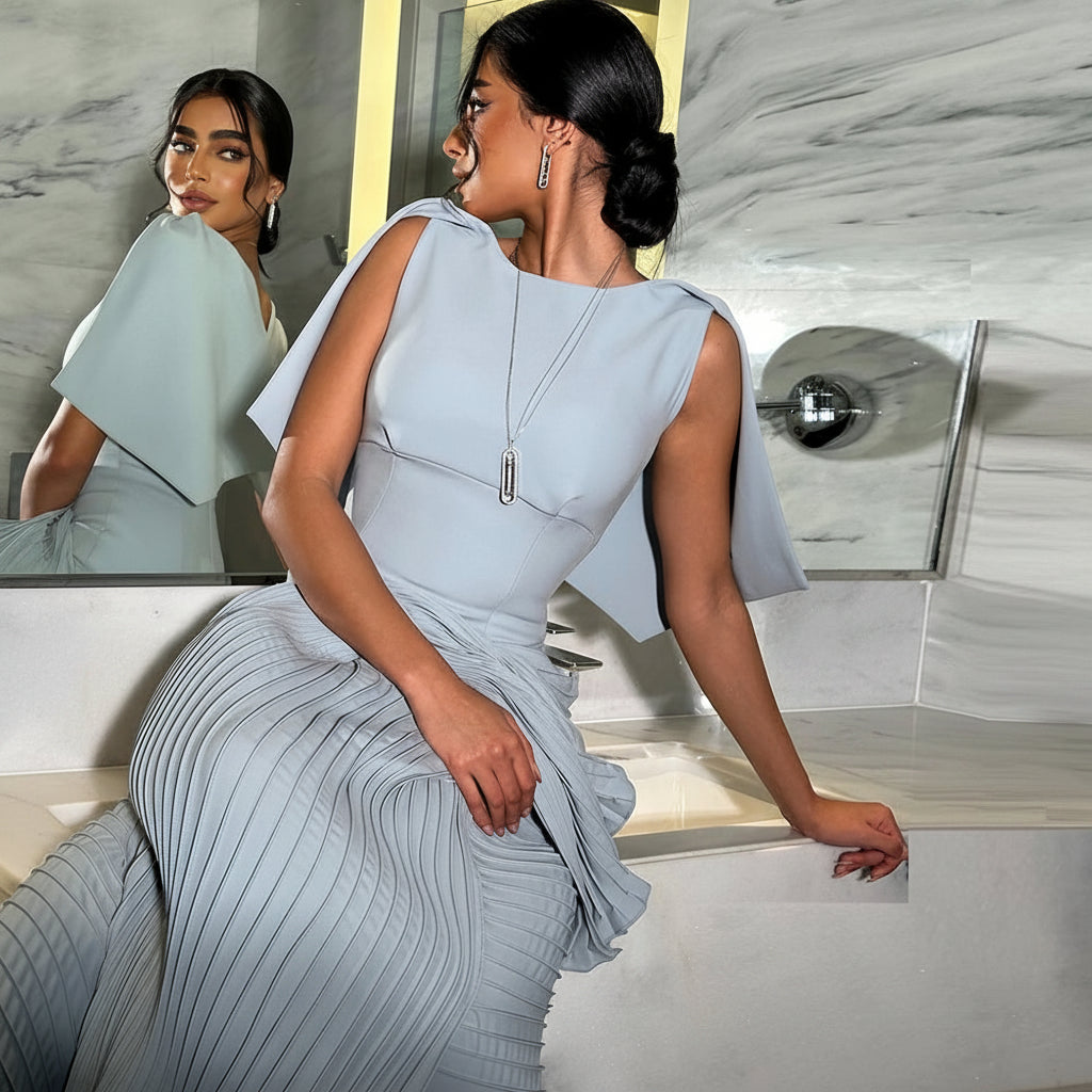 A woman models a light blue pleated dress with a structured bodice and draped cape-like sleeves in a modern bathroom setting, with a mirror reflecting her profile. The text 'Vogue Dress Store' is overlaid at the bottom.