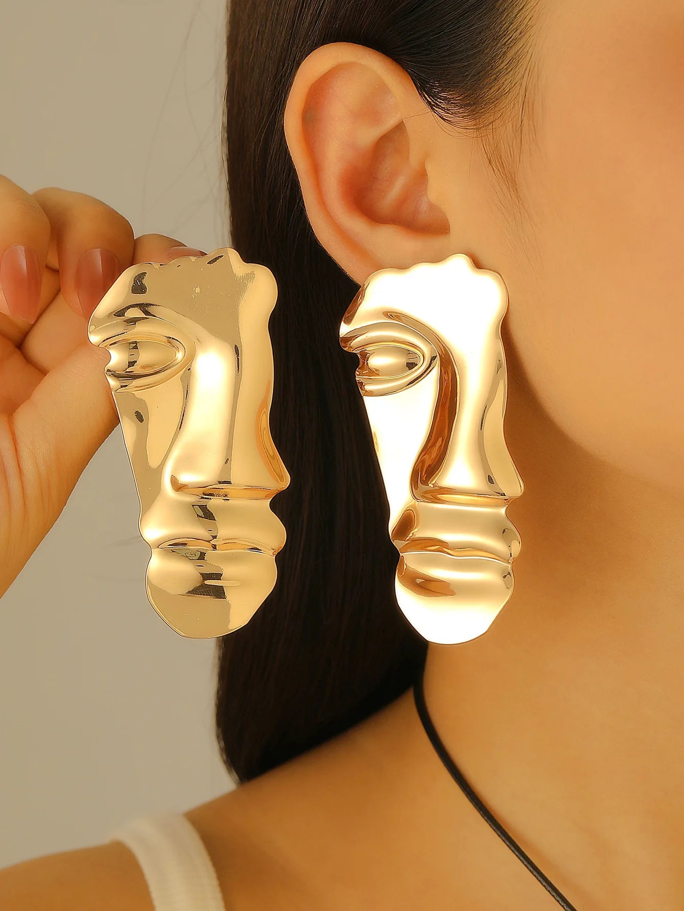 Gold face-shaped earrings worn by a person with a neutral background