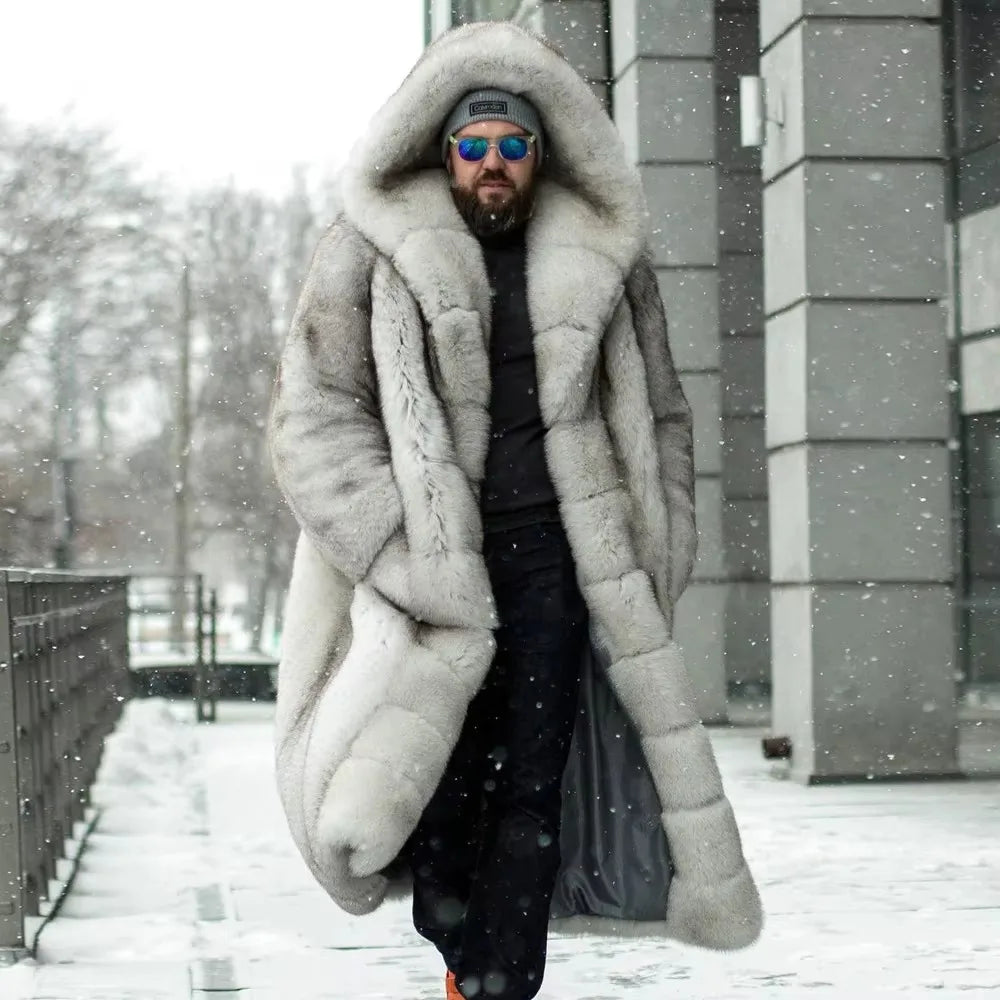A man wearing a long, oversized light gray fur coat with a large hood, black clothing, a gray beanie, and blue-tinted sunglasses, walking outdoors in the snow in front of a modern gray building.
