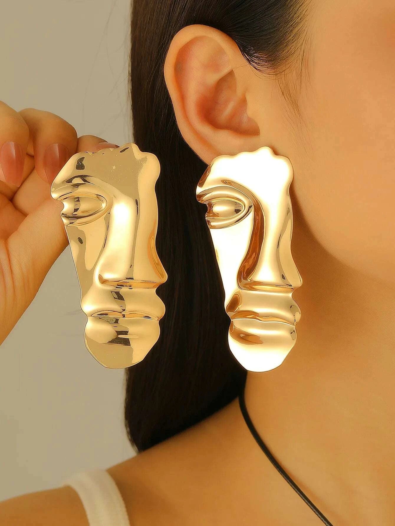 Gold face-shaped earrings worn by a person with a neutral background