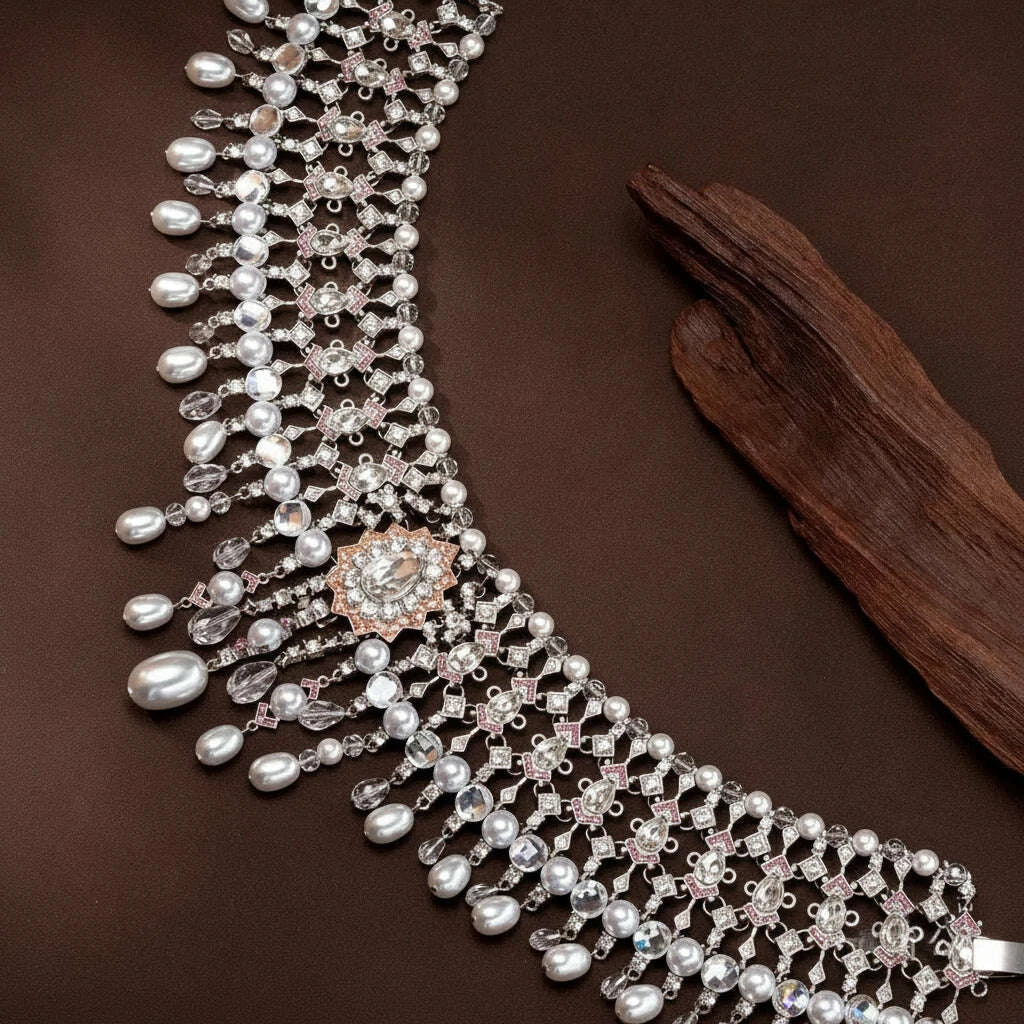 An ornate silver-toned necklace with dangling faux pearls, clear faceted crystals, and a rose-gold-toned floral centerpiece, displayed on a dark brown surface next to a piece of driftwood. Chinese text is visible in the upper left corner.