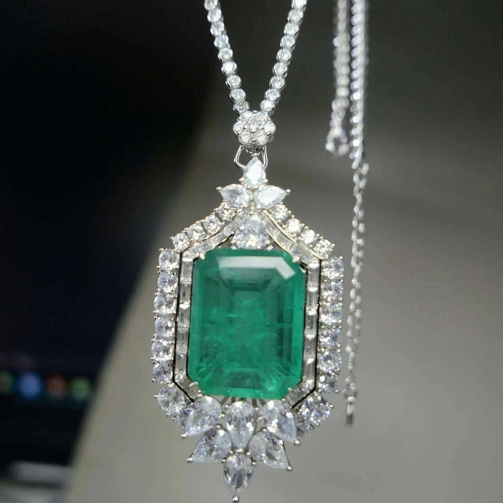 A close-up of a silver pendant necklace featuring a large emerald-cut green gemstone surrounded by clear stones, hanging against a blurred dark and light background.